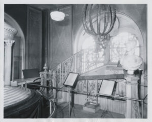 Photograph of interior with a sculpted bust on display in a gallery. In the background, a staircase leads upward with a beautiful, intricate banister.