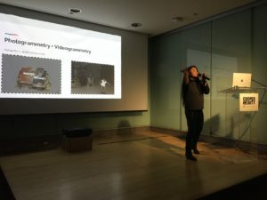 A petite Asian woman with long hair is standing on stage, speaking into a microphone and gesturing with her right hand. Behind her is a screen showing a slide titled: "Photogrammetry + Videogrammetry". To her left is a podium with an open computer on it. Beneath the computer there is a sign that shows the Cooper Hewitt logo.