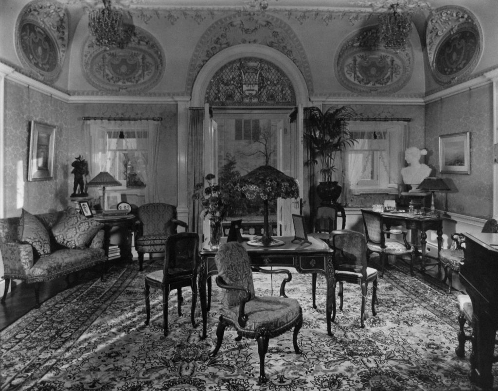 Black and white photo of medium sized room with high ceilings. Sofas, chairs, and a desk furnish the room and a door on the back wall opens to a garden.