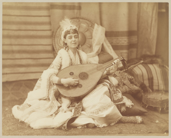 Woman laying down on a pile of pillows in lots of layered, drapey clothing. Sword on the ground to her left and a peacock feather fan beside it.