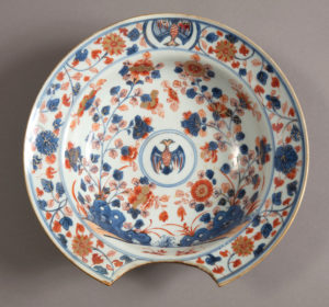 Circular white porcelain shaving basin with a semi-circular section cut out at the bottom, elaborately decorated with blue and red enamel dahlia-like flowers and curling foliage accentuated with gold leaf, a double-headed bird sits in a blue-rimmed circle in the central bowl