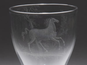 Close-up photograph of the vessel portion of a drinking glass with an image of a horse engraved into its surface.
