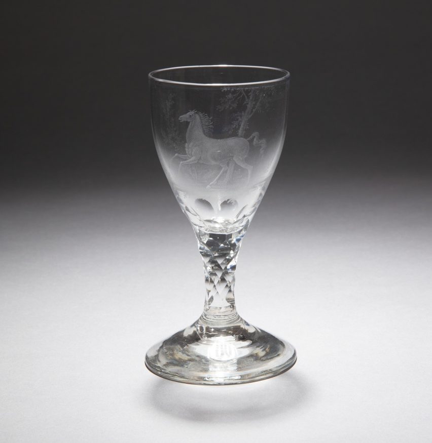 Clear glass with a wide-rimmed ovoid bowl, thick faceted stem, and a conical base. A translucent galloping horse set against thin leafy trees is etched into the surface of the bowl.