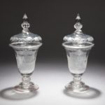Pair of covered clear glass, inverted-bell shape drinking vessels, with knopped stems, stepped circular feet, and squat dome-shaped removable lids with baluster finials. The bodies etched with scenes of female domestic life after the birth of a newborn; the lids etched with garlands.