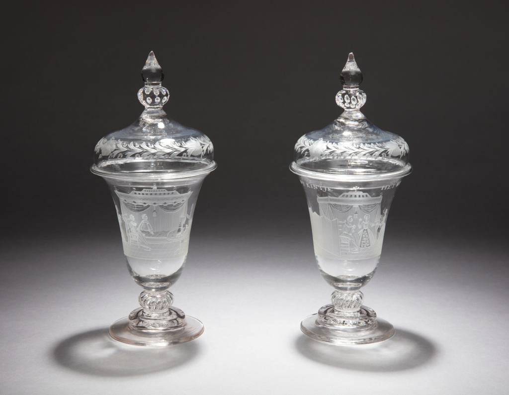 Pair of covered clear glass, inverted-bell shape drinking vessels, with knopped stems, stepped circular feet, and squat dome-shaped removable lids with baluster finials. The bodies etched with scenes of female domestic life after the birth of a newborn; the lids etched with garlands.