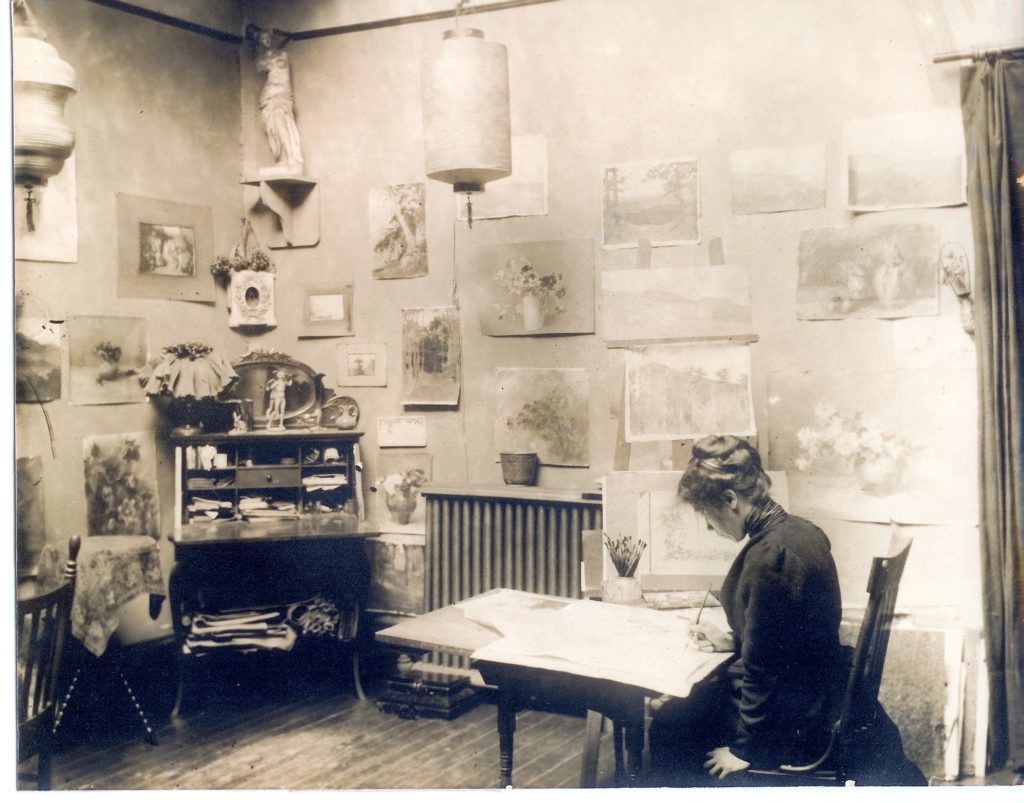 A black and white digital print of Sophia Crownfield sitting at a desk bent over her design in her work room with prints hung on the back wall.