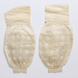 A pair of cream linen and cotton undersleeves with a design of repeating diamonds some appearing embossed others formed from cut-out circles along a puffed portion that tapers into a smaller cuff with a cutout design and a row of flowers along the edge