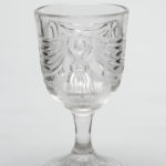 Clear glass goblet with a wide-rimmed ovoid bowl, a round fringed tassel with sweeping swags on either side is pressed into the center of the bowl, the bowl tapers into a thin, conical stem that extends to a faceted round foot.
