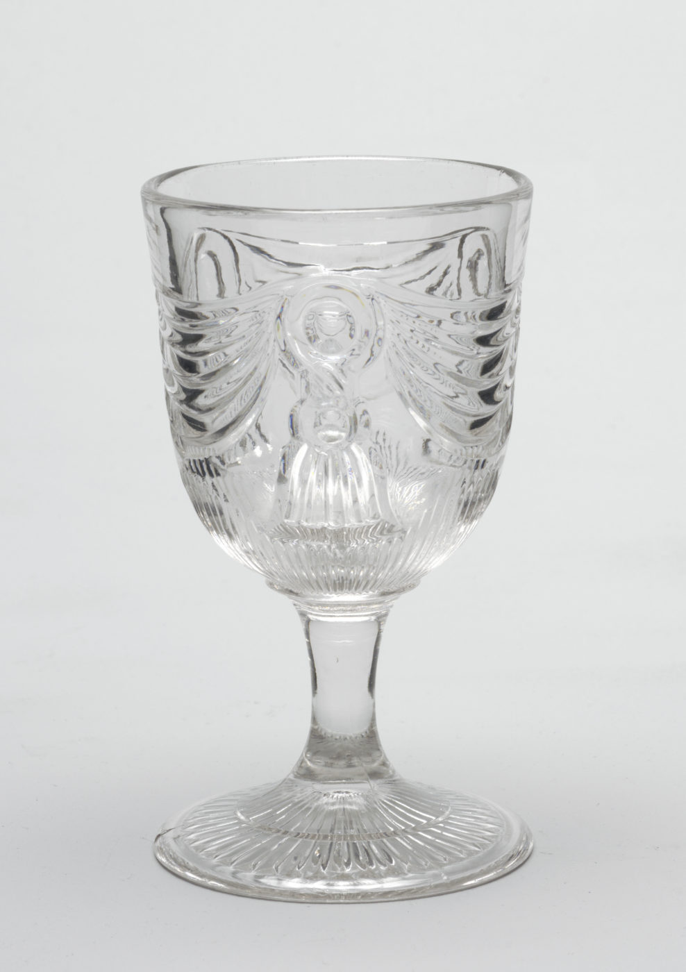 Clear glass goblet with a wide-rimmed ovoid bowl, a round fringed tassel with sweeping swags on either side is pressed into the center of the bowl, the bowl tapers into a thin, conical stem that extends to a faceted round foot.