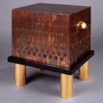 Four short golden cylinders support a flat black platform, on top of which rests a large copper cube with rows of coins affixed to the sides.