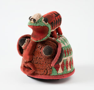 Beaded, dome-shaped, ceremonial hat made up of a rust-colored stylized face, with a patterned cream, red and green helmet-like covering, on top of which stands a chameleon-like creature with a red patterned body and a green face whose mouth is gaping