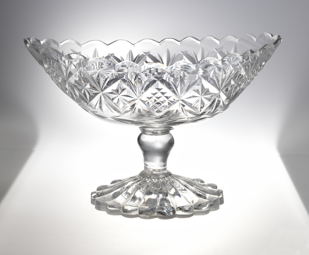 An oval-shaped, footed glass bowl cut with intricate geometric designs and a scalloped rim.