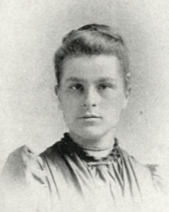 Black and white portrait of a woman with her hair in a bun, wearing a dark gray dress and looking directly at the viewer.