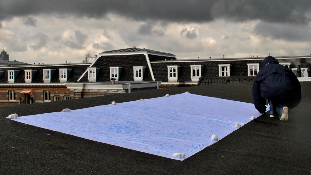 Photo of a person in a black raincoat kneeling on the ground next to a large rectangular sheet spattered in blue and weighed down by fist-sized rocks evenly spaced along its edges.