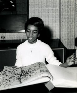Black and white photo of Althea McNish, a dark-skinned woman with short black hair looking through a large book of textile patterns.