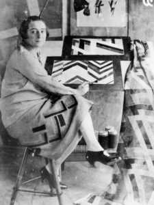 Faded, black and white photo of Sonia Delaunay, a light-skinned woman whose dark hair is cropped at her cheekbones, seated with her legs crossed on a stool in front of a work station on which sit a few geometrically patterned artworks.