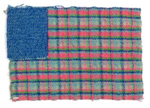 A rectangular woven textile reminiscent of the American flag, with a solid blue rectangle in the upper left corner where the stars usually are and a blue, pink, and green plaid pattern where the stripes would be.