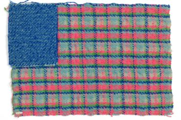 A rectangular woven textile reminiscent of the American flag, with a solid blue rectangle in the upper left corner where the stars usually are and a blue, pink, and green plaid pattern where the stripes would be.