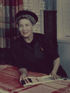 Color photograph of a woman wearing a black hat with a black suit with a pencil in breast pocket, leaning over a table while holding fabric swatches in her hands