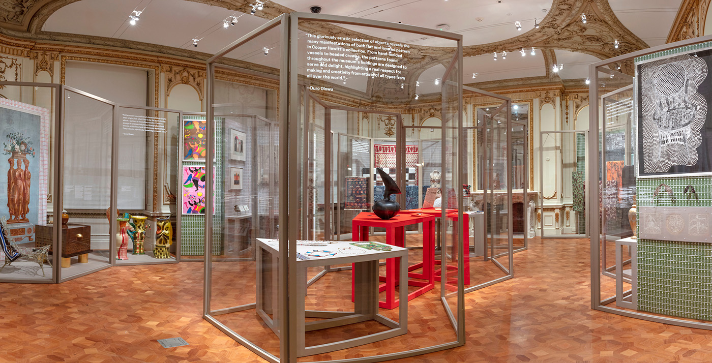 Interior installation photograph inside a room with intricate carved molding. Objects displayed behind a series of tall folding screens.