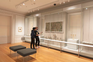 Installation photograph inside a white room with light-wood floors, two figures observe objects behind plexiglass, including an intricately-patterned wallcovering, silver baskets and tea caddies flanked by golden candelabras. To the left a textile with plates beneath, to the right four clear goblets, two grey cushioned stools sit in foreground.