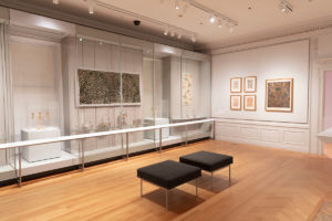 Installation photograph inside a white room with light-wood floors, objects displayed behind plexiglass from left to right include a gold candelabra, silver baskets and tea caddies, a second golden candelabra, and an intricately-patterned wallcovering. Further to the right is a textile with plates beneath, and outside of the glass hanging on the wall are small brightly patterned designs framed in gold. Two grey cushioned stools sit in foreground.