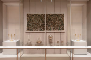Close-up photograph of a section of objects displayed behind plexiglass in a white gallery. In the center of the glass display case is a long, horizontal-rectangular cream canvas mounted on the wall with an intricate olive-green repeating design of leaves and flowers with accents of orange and light green. Underneath is a row of silver objects placed on a clear glass shelf, including two shallow silver baskets with their handles up on the left, a silver tea urn in the shape of Atlas supporting the world in the center, and two elaborately-decorated cylindrical tea caddies on the right. On either side, also behind glass, are two recessed windows with cream blinds drawn down, in front of them are two white platforms, on each stands a golden three pronged candelabra.
