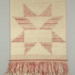 Rough-looking textile made of woven light tan strands, with red strands forming an abstract geometric star shape in the center; a long fringe of red and tan strands hangs along the bottom.