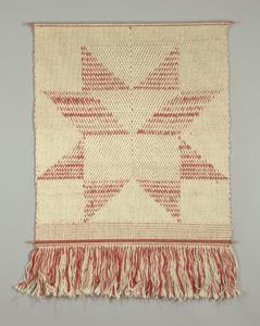 Rough-looking textile made of woven light tan strands, with red strands forming an abstract geometric star shape in the center; a long fringe of red and tan strands hangs along the bottom.