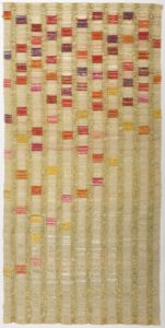 Vertical columns of light golden thread run down the length of a long textile, with vibrant squares of pink, purple, red, and yellow cascading down the columns.
