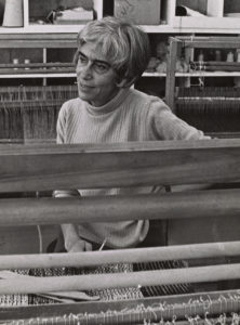 Black and white photo of Trude Guermonprez, a woman with light skin, thick, dark eyebrows, and short salt-and-pepper hair; she is seated amongst various textile looms.