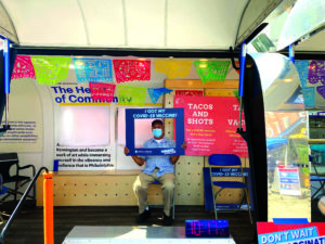 A man sits on a chair inside a van with an open side. He is holding a cardboard frame over his face labeled, "I got my COVID-19 vaccine!" The van is full of colorful posters. They read, "Don't wait, vaccinate!" and "Tacos for Shots."
