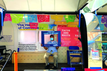 A man sits on a chair inside a van with an open side. He is holding a cardboard frame over his face labeled, "I got my COVID-19 vaccine!" The van is full of colorful posters. They read, "Don't wait, vaccinate!" and "Tacos for Shots."