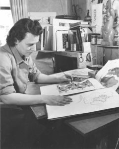 Black and white photo of Lanette Scheeline, a woman with light skin and dark, shoulder-length hair, holding a thin paintbrush with several pieces of floral art laid out in front of her.