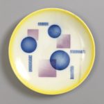 A round, cream-colored plate with a bright yellow edge and blue and purple circles, rectangles, and squiggly lines scattered throughout the center.
