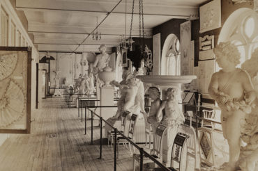 Faded photograph of a gallery space that has rows of chairs and sculptures of cherubs and urns on the right, on the left is a paneled storage wall with a panel containing two fans pulled out.