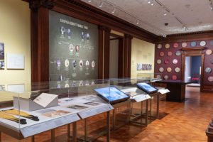 A museum gallery with cases of objects and wall graphics of a family tree and a textile pattern.