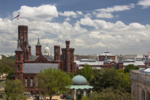 Sunny exterior view of the Smithsonian Castle, in brown brick with numerous towers, against a blue sky with fluffy white clouds.