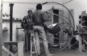 Two men stand on either side of a sculpture of concentric metal circles in what looks like an artist studio with worktables and tools.