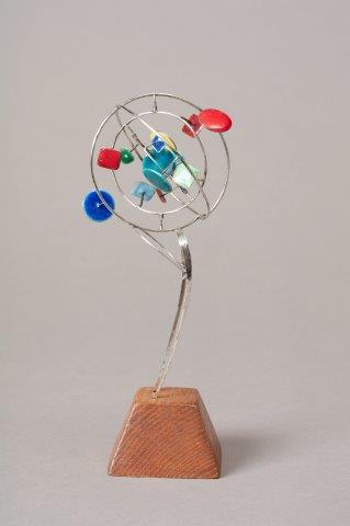 A sculpture of metal wire and color shapes sits on a single slender stem that rests on a wooden pedestal.