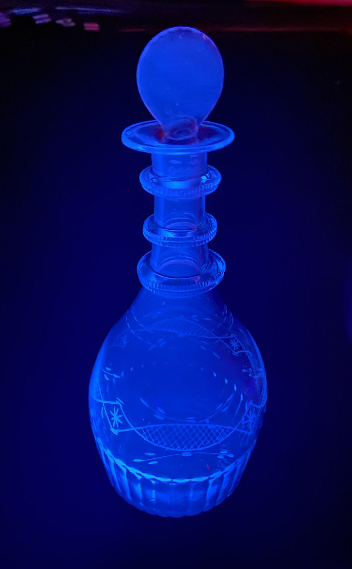 A glass decanter, with a bulbous bottom ascending into a narrow-necked top and a disc-like stopper, glows electric blue in a dark space.