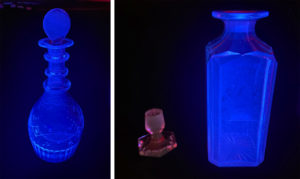 Two glass decanters glow electric blue in a dark black space with a decanter stopper, lit faintly pink, stationed between them.