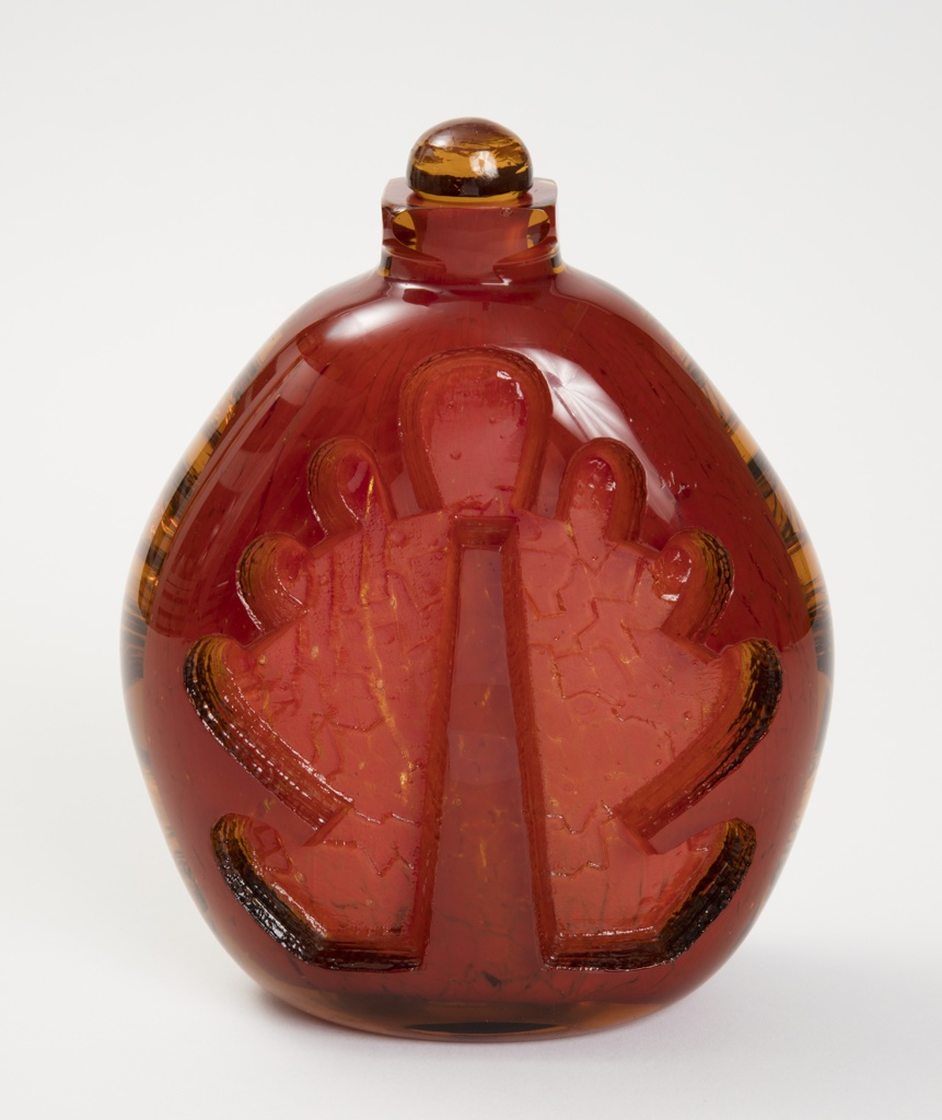 Stout brick-red glass vessel with amber details on its sides and amber half round removable stopper resting on its short cylindrical neck.
