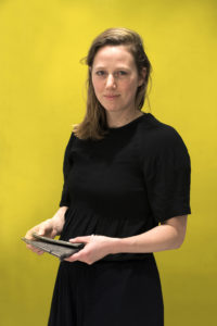 Color photograph of a woman with long hair, looking directly at the camera, holding a bag in her hands while standing against a yellow background.