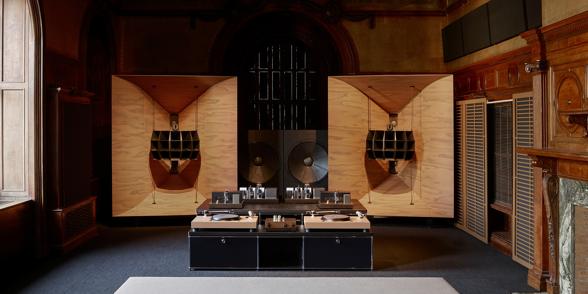 Interior view of a large, high-end audio system, including speakers, subwoofer, and turntable setup. with details in light-colored wood. The system is set up in a room with ornate wood architecture details and a wide window.