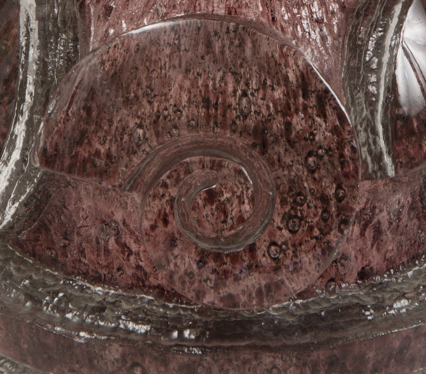 Detail of a purple glass object highlighting the raised, spiraled form of a snail shell with a bubbled texture in the material of the glass.