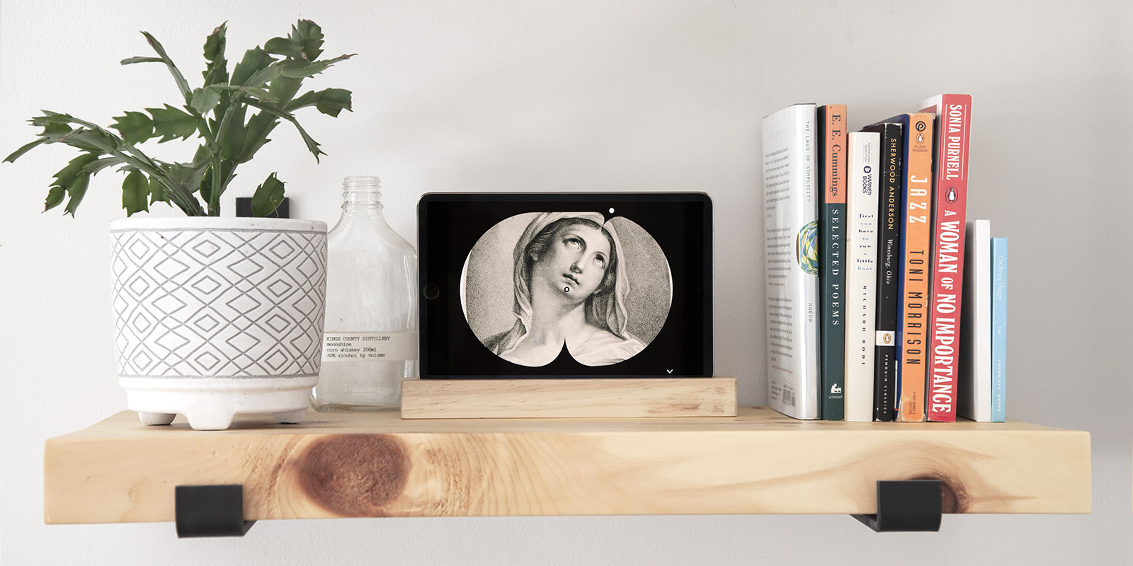 The finished Art Clock displayed on a horizontal tablet sitting on a light wood shelf with books to the right and a Christmas cactus to the left. The Art Clock shows a close cropped etching of a medieval figure with the minute hand pointed to their chin (at 30 past the hour) and the hour hand a 1pm, corresponding to the angle of the figure's eyes.