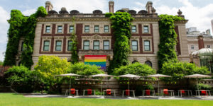 A photograph of a large brick and stone building that is set against a blue sky and a field of green grass stretched before it. Hung from the middle of the building is a rectangular rainbow flag, composed of six stripes of red, orange, yellow, green, blue, and purple.