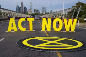 Bold, yellow letters spelling out [Act Now] standing in middle of highway with large yellow circle with x-shape flat on the ground in front. Skyline and demonstrators in background