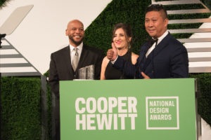 Three people stand triumphantly behind a podium at the National Design Awards. One has a hand in the "thumbs up" pose and a glass NDA trophy rests on the podium.